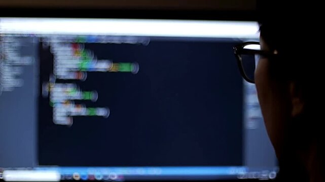Over-the-shoulder shot of a programmer or hacker coding on a computer screen in a dark room.