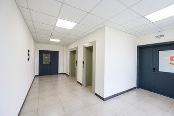 brightly lit hallway features neutral walls, tiled floors, and a suspended ceiling. Two elevators stand between doors. The number "5" is on a wall
