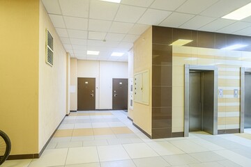 well-lit corridor with tiled floors, brown doors, and elevator access. The walls are painted in shades of yellow and beige