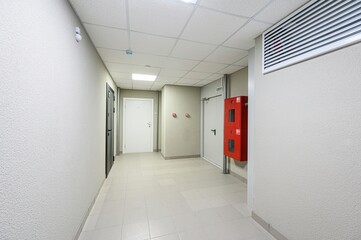brightly lit hallway with tiled floors. It features doors, a red fire extinguisher box, and a textured wall. The ceiling has square panels