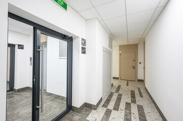 Bright hallway with glass doors, white walls, and geometric patterned flooring. Exit sign visible