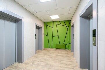 modern hallway with three grey elevators, a green accent wall with black geometric design, light wood-look flooring, and a drop ceiling with fluorescent lighting