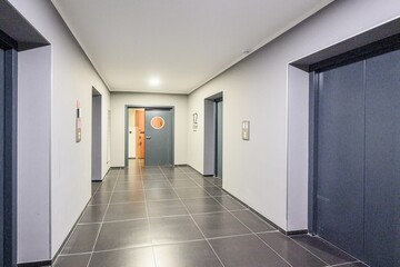 modern hallway with tiled floors, grey walls, and elevator doors. There's a door with a round...