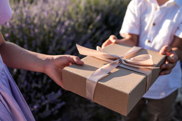 Close-up of two children exchanging a kraft gift box with a ribbon and a lavender bouquet in a...