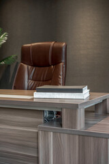 Closeup of a wooden office desk with books, with a luxurious brown leather executive chair blurred in the background.