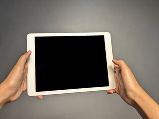 Two hands holding a white tablet computer with a black screen against a plain gray background in a close-up shot