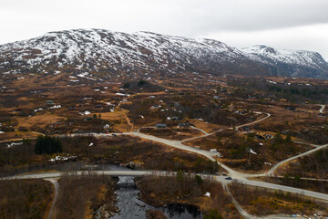 Dramatic terrain surrounding Voringsfossen waterfall against snow-capped mountain. Winding road and flowing river cut through dark rugged landscape