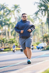Obraz premium An overweight man jogging on a street with traffic in a tropical city. Promoting healthy lifestyle, fitness, and motivation. Wearing hoodie and shorts, doing morning exercise