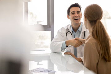 Obraz premium Joyful young Latin physician man shaking hands with female patient, smiling, laughing, congratulating on recovery, checkup perfect results, good news. Candid shot with copy space