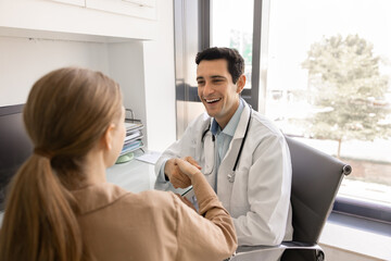 Happy young Hispanic doc giving greeting handshake, medical advice to healthy woman at workplace in...