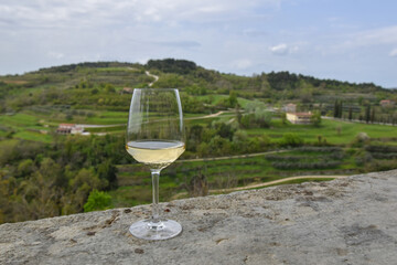 A glass of white wine on a stone ledge with a view of green hills, winding roads, and scattered...