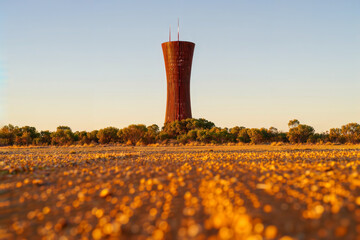 Water tower in the outback. A uniquely shaped, rust-colored water tower stands in a dry, rural landscape under a clear sky at sunrise.