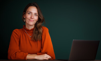 A woman in an orange blouse sits at her desk with a laptop, smiling and looking into the camera against a dark green background. 
