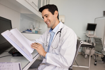 Positive satisfied young Hispanic physician man checking patient history at workplace, thumbing...
