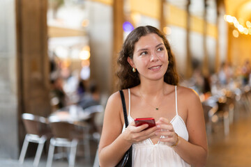 In evening, smiling girl with smartphone walks along city street Placa Real, messenging with friend...