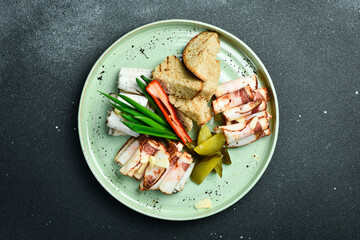 Sliced salted lard with brown bread and vegetables. Ukrainian traditional food. On a plate. Restaurant serving. Close up. On a dark stone background.