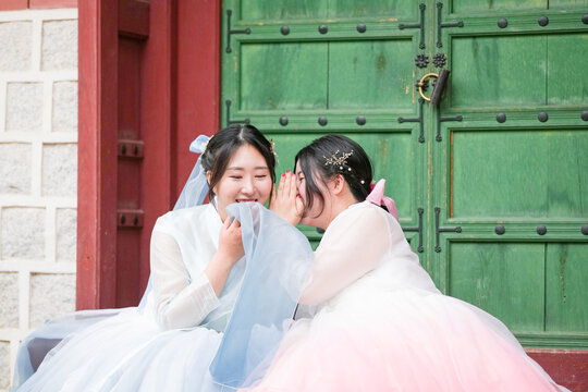 Two Korean women in their twenties sit before the heavy metal door of a historic Gyeongbokgung Palace hall under a clear summer sky, wearing cute pink and sky-blue hanbok while chatting. Seoul, June.