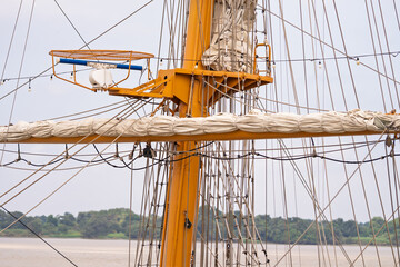 Ship mast with furled sails and visible ropes in a natural riverside setting.