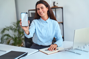 Professional Businesswoman Smiling While Showcasing Smartphone With Blank Screen, Sitting at Office Desk with Laptop and Notebook. Promotes Technology, Communication, and Professionalism