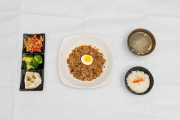 A Thai basil minced pork set meal arranged on a tray, including a portion of stir-fried spicy basil pork, a bowl of steamed white rice, three side vegetables, and a bowl of soup. The presentation refl