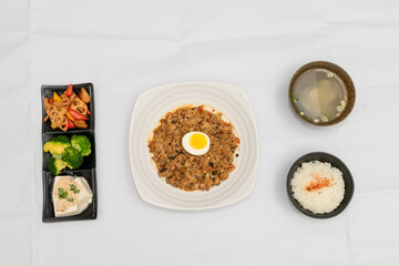 A Thai basil minced pork set meal arranged on a tray, including a portion of stir-fried spicy basil pork, a bowl of steamed white rice, three side vegetables, and a bowl of soup. The presentation refl