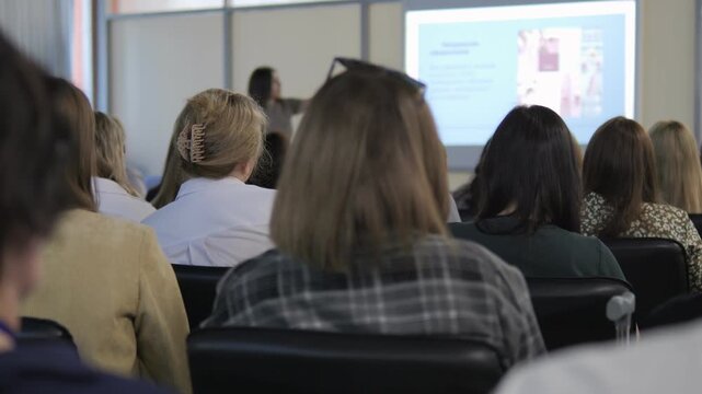 Group of people are sitting in a classroom. A woman is standing in front of a projector screen
