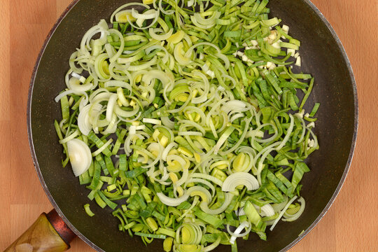A generous amount of freshly sliced green and white leeks is spread evenly in a dark frying pan, resting on a wooden surface