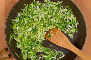 A wooden spoon is stirring sliced green and white leeks in a dark frying pan, positioned on a...