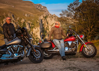 Mature couple posing with two cruiser motorcycles in scenic mountain setting during golden hour, expressing freedom, style, and adventure.