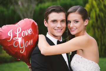Happy young bride and groom hug outdoors, holding an "I Love You" balloon. Smiling couple portrait on their wedding day, showing romance and love.