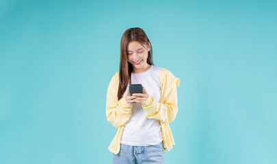 Smiling young Asian woman looking at smartphone while standing against a sky blue background,...