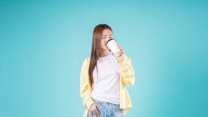 Relaxed young Asian woman enjoying the aroma of coffee while standing against a blue background,...