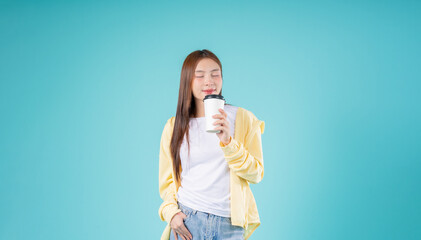 Relaxed young Asian woman enjoying the aroma of coffee while standing against a blue background,...