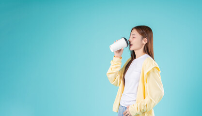 Young Asian woman and coffee cup while standing against a blue background, reflecting modern...