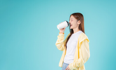 Young Asian woman and coffee cup while standing against a blue background, reflecting modern...