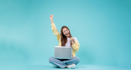 Excited young Asian woman sitting cross-legged with a laptop, raising her hand in celebration...