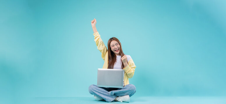Excited young Asian woman sitting cross-legged with a laptop, raising her hand in celebration against a blue background, symbolizing success, online learning, and remote work.