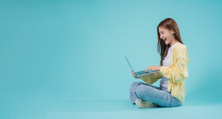Happy young Asian woman sitting cross-legged using a laptop against a blue background, representing...
