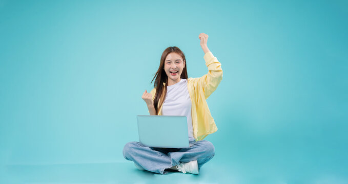 Excited young Asian woman sitting cross-legged with a laptop, raising her hand in celebration against a blue background, symbolizing success, online learning, and remote work.