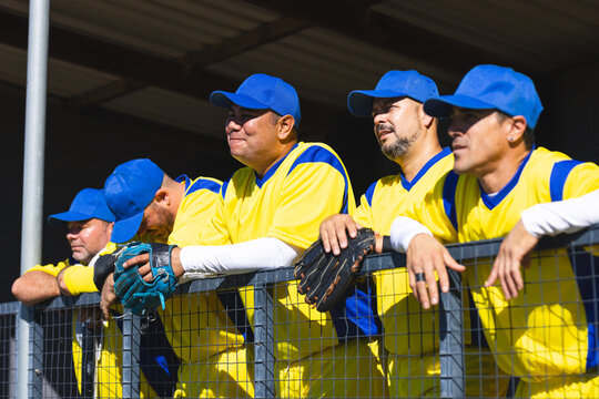Male baseball teammates leaning on fence at baseball dugout in yellow jerseys holding gloves