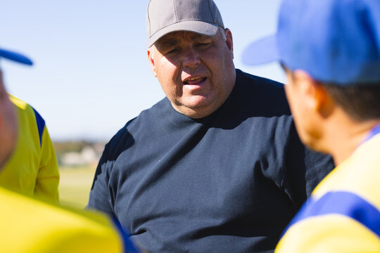 Male coach instructing players in yellow jerseys on grass field wearing baseball cap and navy shirt