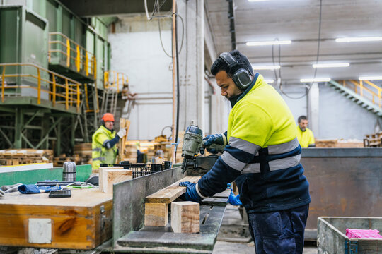 Carpenter wearing safety equipment using pneumatic nail gun assembling wooden pallets in a recycling factory