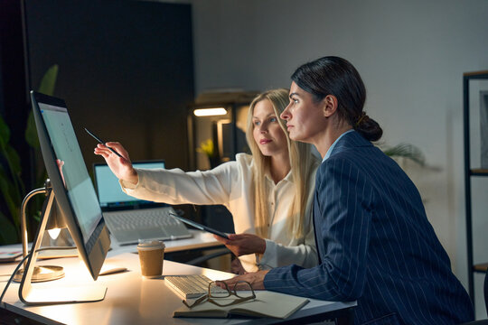 Two Professional Women Collaborating In An Office Setting, Using Desktop Computers And Engaging In Discussion During A Night Work Shift. Modern Workplace, Teamwork, And Business Environment Concept