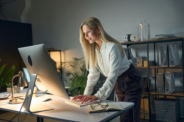 Professional woman working on a desktop computer in a modern office setting late at night,...