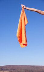 Human hand holding an orange microfiber hiking towel against a clear sky background. Bright outdoor gear for travel, hygiene, and post-shower relaxation