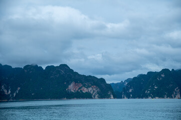 A view of towering limestone mountains in a tranquil lake under a cloudy sky in southern Thailand.
