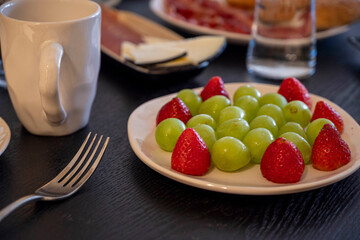 A plate of fruit with green grapes and red strawberries on a table. The plate is on a white plate and is surrounded by a fork and a cup