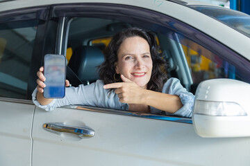 Middle-aged Caucasian woman sitting inside a car, holding and pointing to smartphone with a smile,...