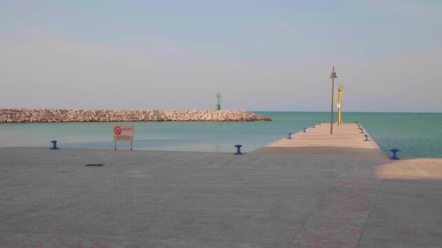 A stone pier leads into tranquil blue sea at Rodi Garganico&rsquo;s port, featuring a green beacon, rocky breakwater, and clear sky&mdash;perfect for maritime themes and coastal serenity.
