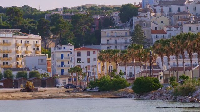 Panoramic view of Rodi Garganico, Italy, featuring a picturesque coastal town nestled against green hills. Palm trees line the shore, with colorful buildings ascending from the sea. Captures Mediterra
